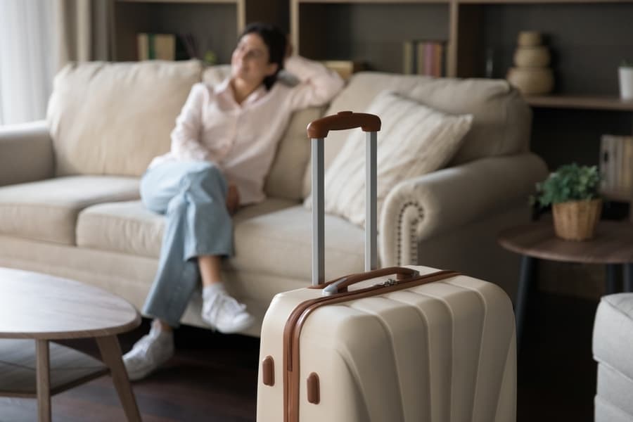 Focus On Beige Suitcase With Blurred Carefree Young Woman Sitting On Sofa On Background