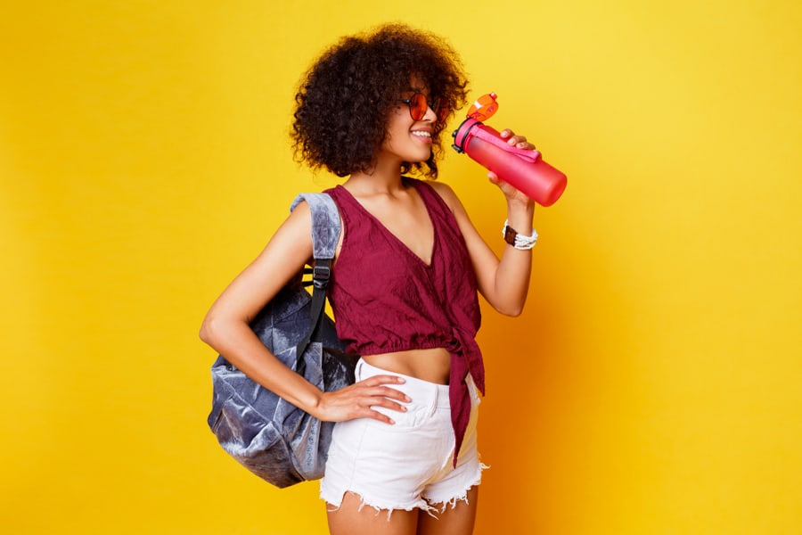 Graceful Sport Black Female Standing Over Yellow Background And Holding Pink Bottle Of Water.