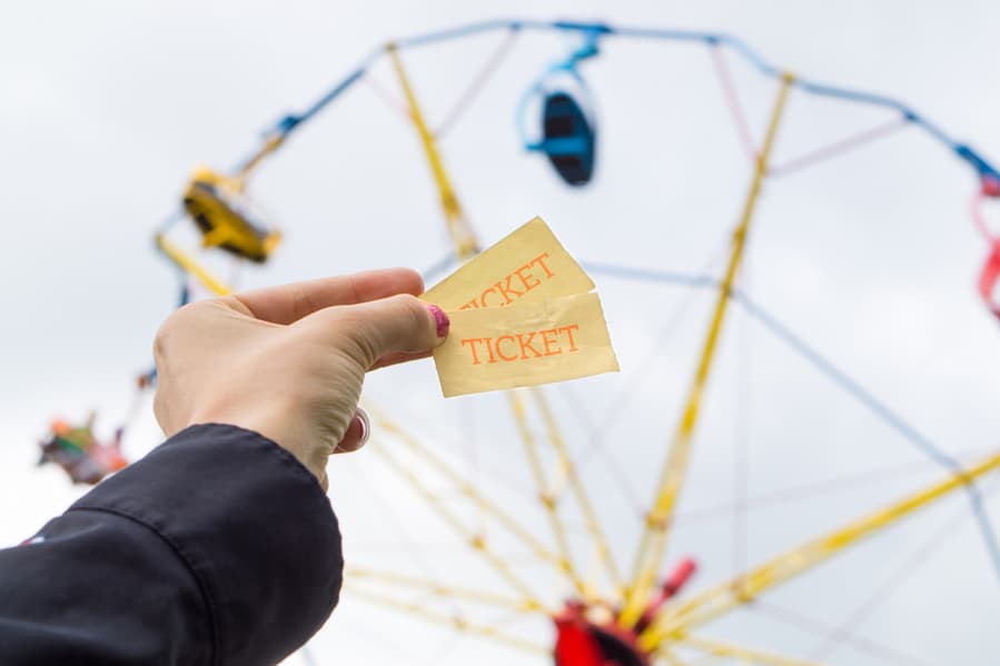 Girl Or Young Woman Holding Amusement Park Tickets In Hand. Colorful Theme Park Ride In The Background.