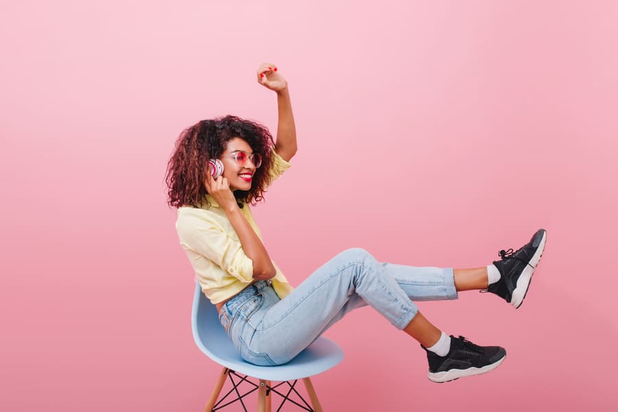 Enthusiastic Black Woman With Curly Hairstyle And In Black Sneaker, Posing In Studio With Blue Interior.