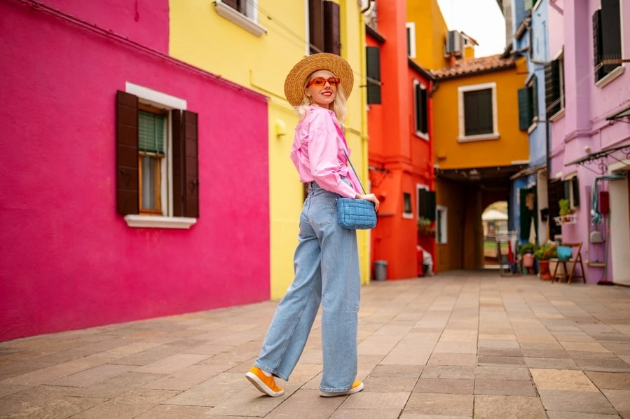 Happy Smiling Female Traveler Wearing Stylish Hat, Glasses, Pink Shirt, Wide Leg Trousers, Walking, Posing Near Colorful Houses In Street.