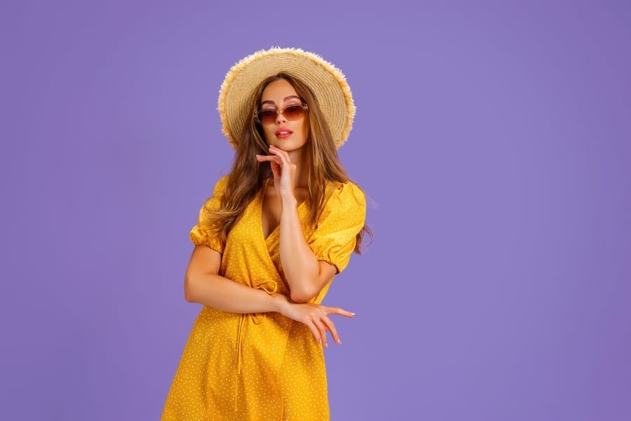 Portrait Of Excited Young Woman In Yellow Dress, Summer Hat, Sunglasses Posing Isolated On Pastel Violet Background.