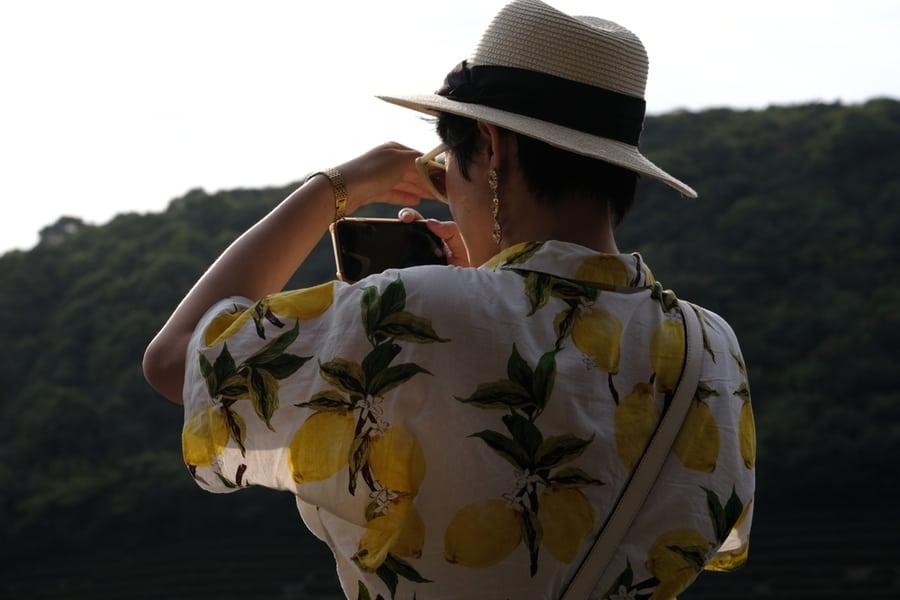 Rear View Of A Short Hair Asian Woman In Straw Hat And Short Sleeve Shirt Using Cellphone Taking Pictures Standing On An Observation Deck. Summer Vacation Concept.