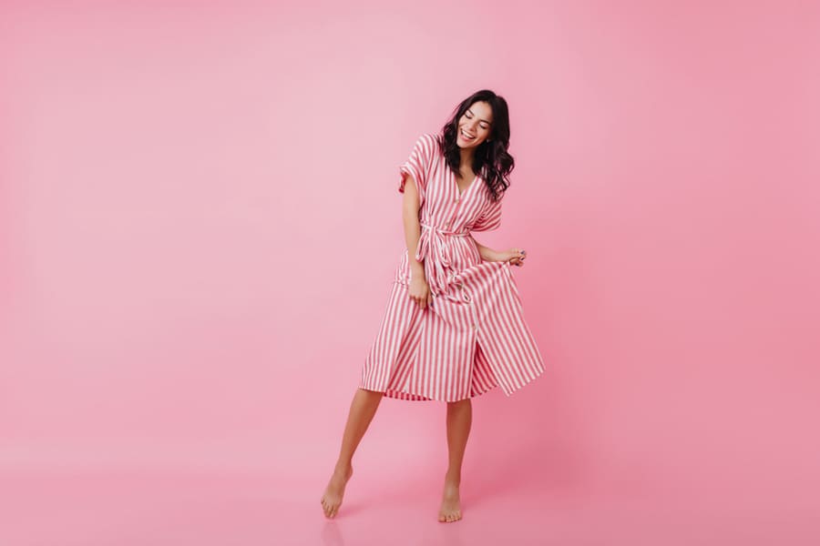 Shapely Barefooted Lady With Tanned Skin Dancing On Pink Background. Happy Caucasian Girl In Striped Dress Fooling Around And Laughing.
