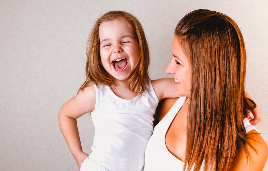 Smiling Young Casual Redhead Female Holding Laughing Little Daughter In White Tank Top While Having Fun Together Standing Against Light Background