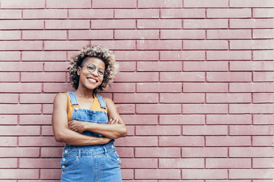 Stylish Smiling African American Woman With Crossed Arms Wearing Casual Bib Jeans Overall Over A Brick Wall