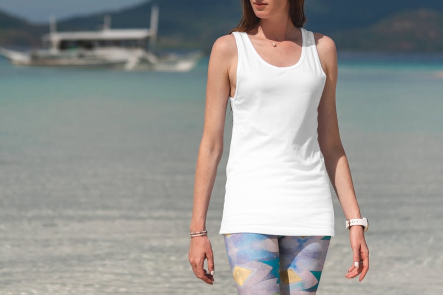 White Tank Top T-Shirt On A Young Woman In Leggins, Front View Mockup, On A Beautiful Beach