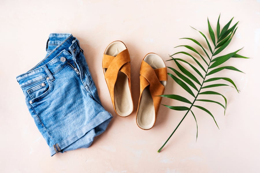 Woman's Casual Trendy Leather Sandals With Crisscross Details And Denim Shorts For Summer Vacation Outfits On Pink Pastel Background With Palm Leaf.