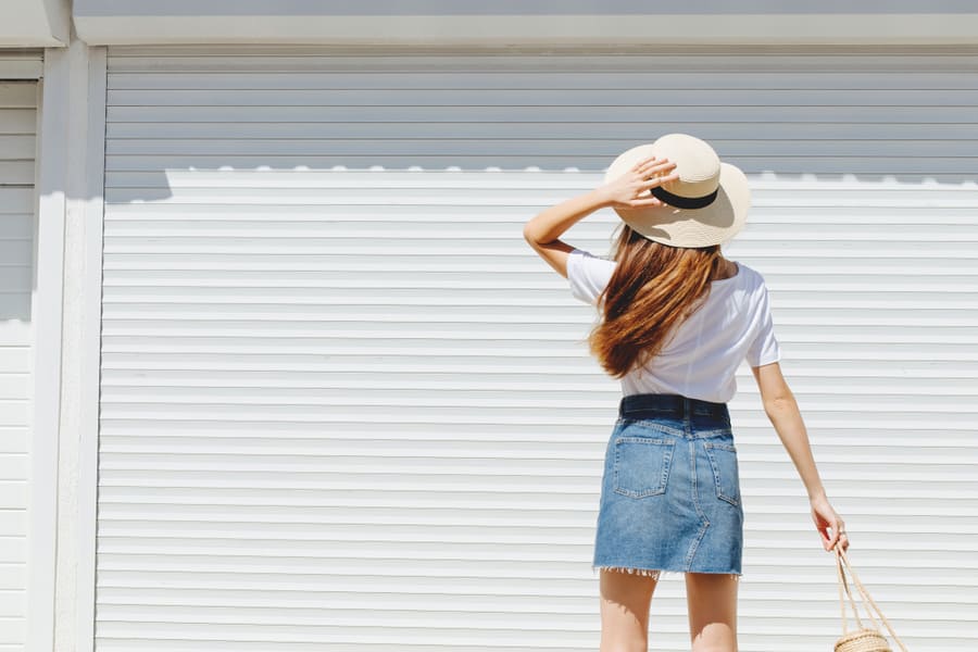 Young Brunette Woman Wearing Blue Denim Mini Skirt, White T-Shirt And Boater Hat Walking Near White Roller Door. Trendy Casual Summer Or Spring Outfit.