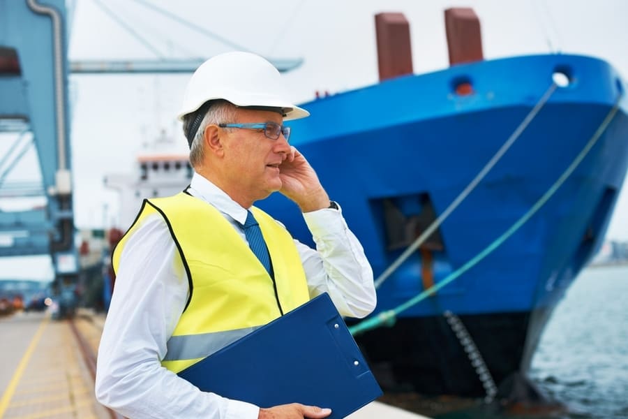 A Dock Worker Standing At The Harbor Amidst Shipping Industry Activity While Taking A Call On His Mobile