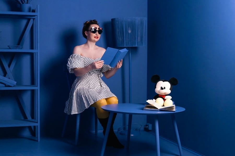 A Woman Dressed In A Black And White Polka Dot Dress And A Black Round Sunglasses Sitting In A Room With A Mickey Mouse Plush Toy Where Everything Is Blue.