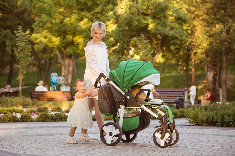 A Young Single Mother In A White Dress, In A Park With Her Daughter, A Girl, In A Carriage, Walking