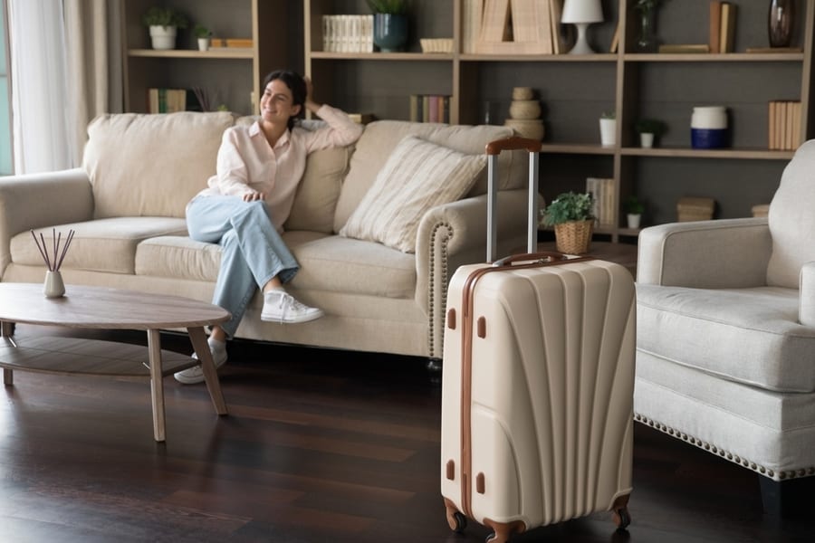 Big Beige Suitcase Standing On Wooden Floor With Woman Sitting On Comfortable Couch