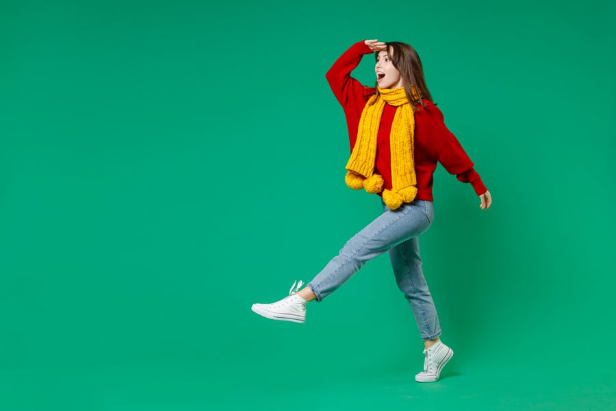 Full Length Side View Of Excited Shocked Young Brunette Woman 20S In Knitted Red Sweater Yellow Scarf Holding Hand At Forehead Looking Far Away Distance Isolated On Green Background Studio Portrait