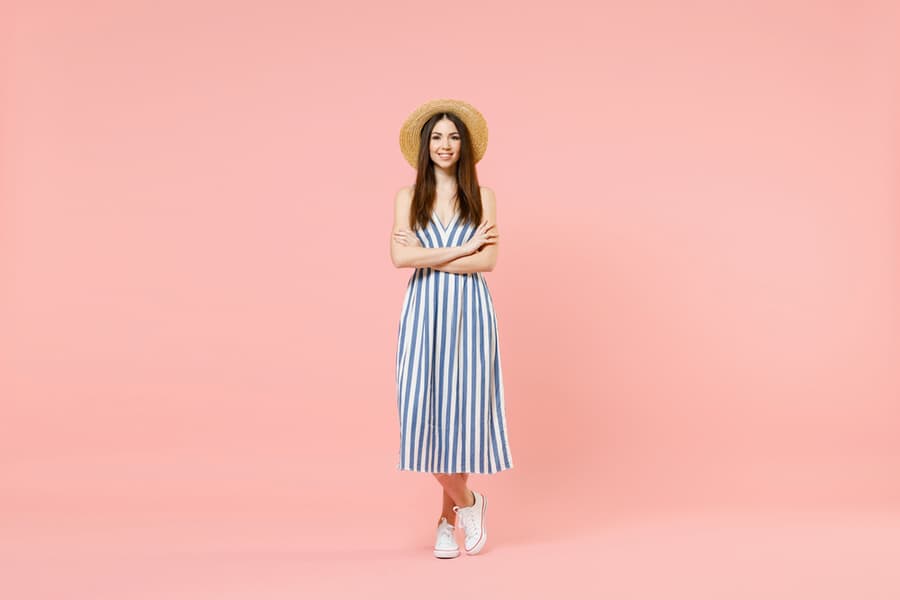 Full Length Young Fun Smiling Cheerful Caucasian Woman In Summer Clothes Striped Dress Straw Hat Hold Hands Crossed Folded Isolated On Pastel Pink Background Studio Portrait.