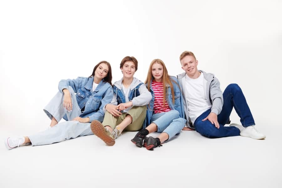 Joyful And Smiling Group Of Teenagers In Trendy Denim Clothes Sitting On A White Studio Background